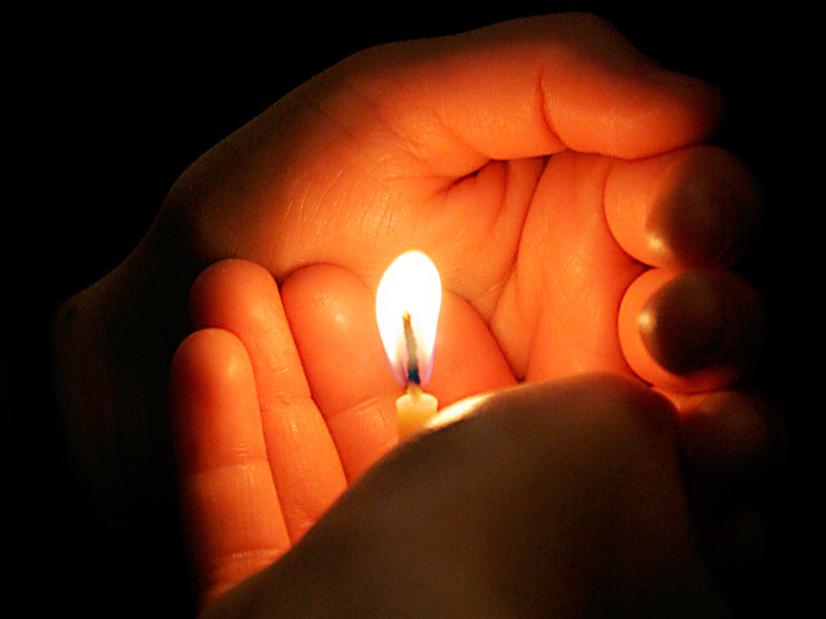 Zwei Hände halten eine Kerze, deren Flamme in der Dunkelheit leuchtet.